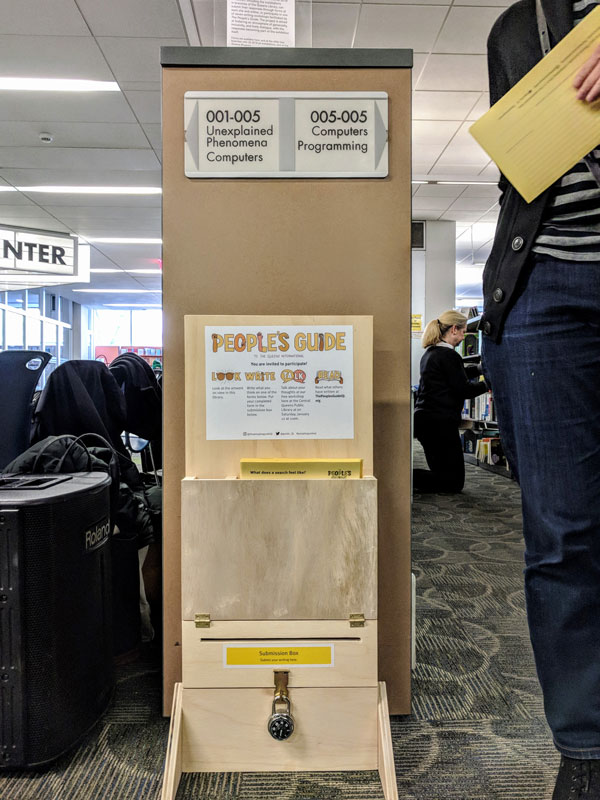 Wooden display holding forms and a submission box, located at the end of stacks of library books.