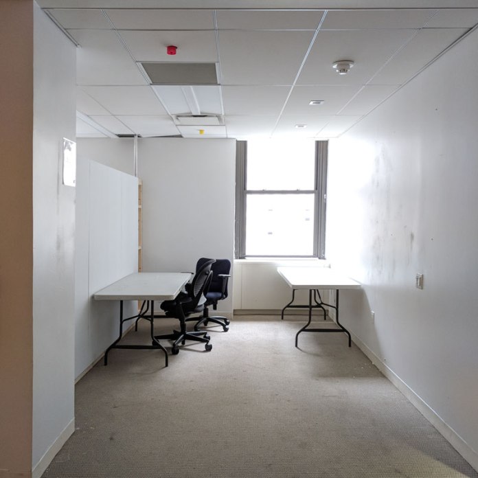A empty white walled room with a bright window in the back corner. Two tables and two office chairs. light brown carpet.