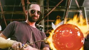 A man wearing sunglasses using a gas torch on a molten ball of glass.